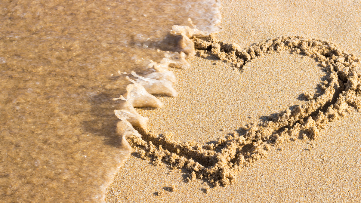 Ocean wave washing over a heart drawn in the sand, symbolizing fading love or letting go at the beach.