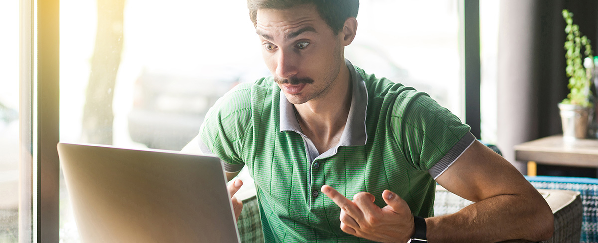 Man in a green shirt sitting at a laptop with a surprised expression, gesturing in frustration at the screen.