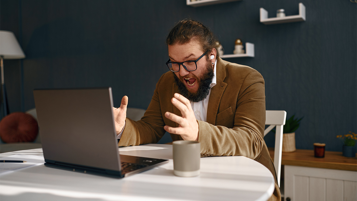 Man in glasses and brown blazer shouting in frustration at his laptop while working at a desk with a coffee mug.