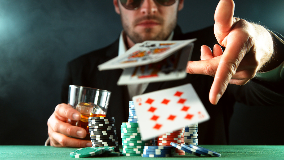 Man in sunglasses tossing playing cards at a poker table, surrounded by poker chips and holding a glass of whiskey, symbolizing risk and decision-making.
