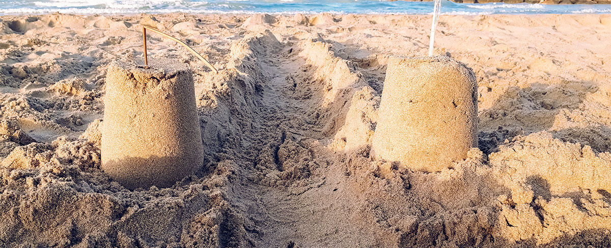 Two small sandcastles with sticks on top stand beside a trench dug in the sand near the ocean shore.
